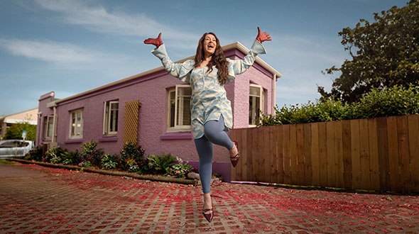 Alyx is dancing with arms in the air in a residential driveway. A pink house in the background.